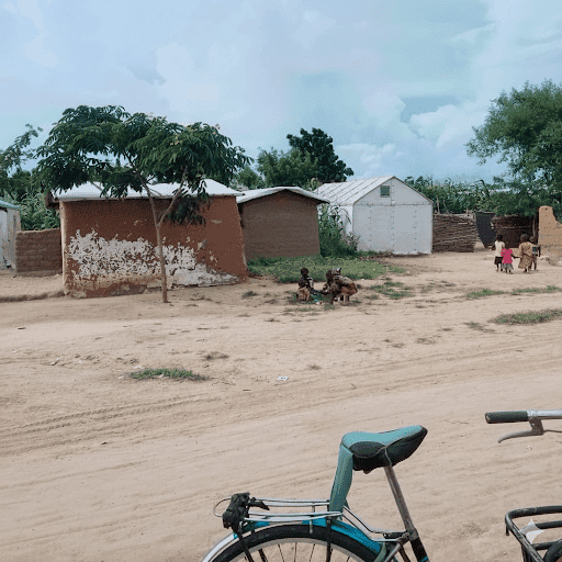 Dirt road with a bicycle in the foreground, houses, trees, and people gathered under a tree in the background.