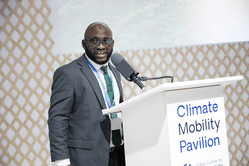 Person speaking at a podium labeled "Climate Mobility Pavilion" with a patterned backdrop.
