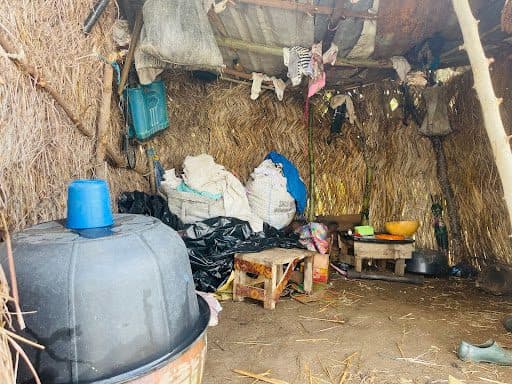 Inside a rustic hut with straw walls, plastic barrels, sacks, and small wooden stools on the dirt floor. Hanging items on the walls.