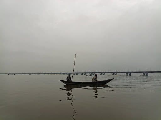 Two individuals in a wooden boat on a calm river, with a long bridge in the background under an overcast sky.