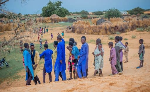 A group of children in colorful clothing stand on a sandy hill, with thatched huts and trees in the background.