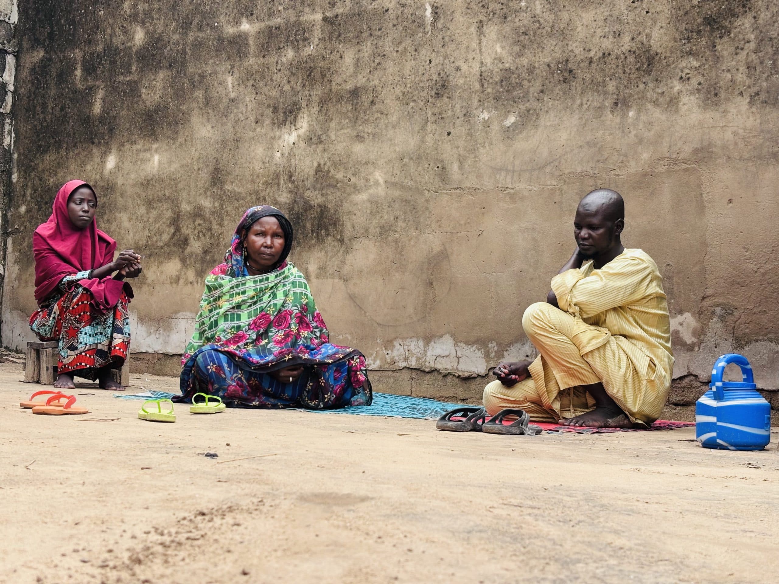 Three people in colorful attire sit against a textured wall, with sandals and a blue container nearby.