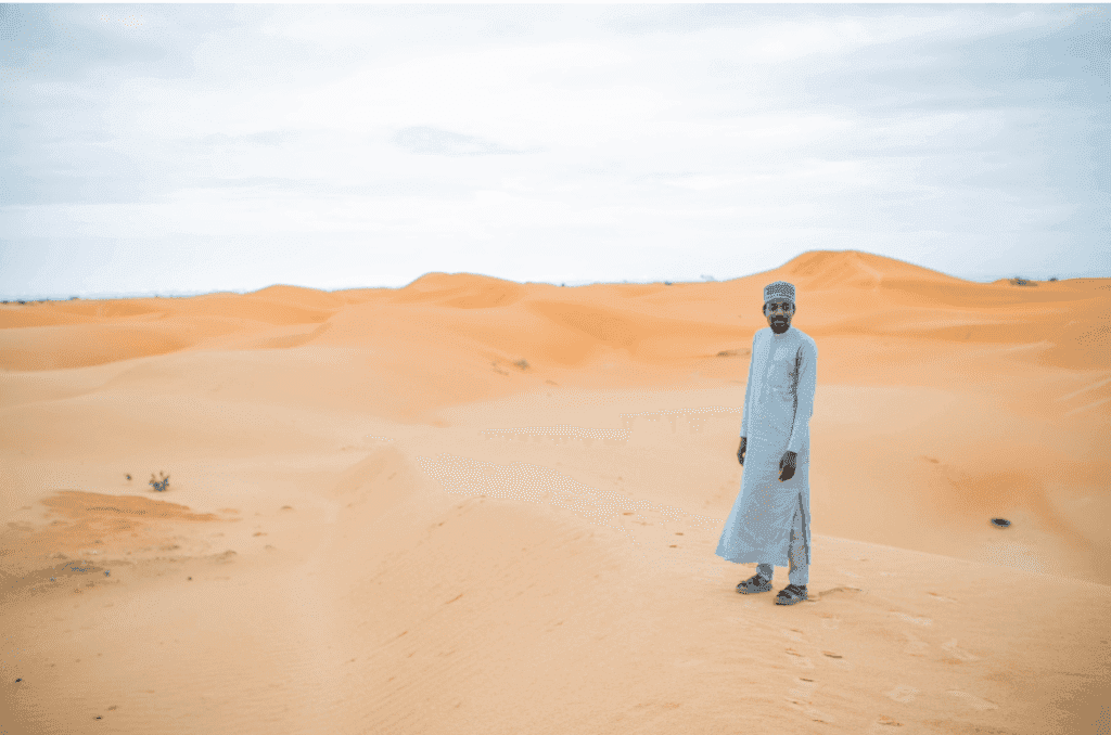 Person in traditional attire standing on sandy desert dunes under a cloudy sky.