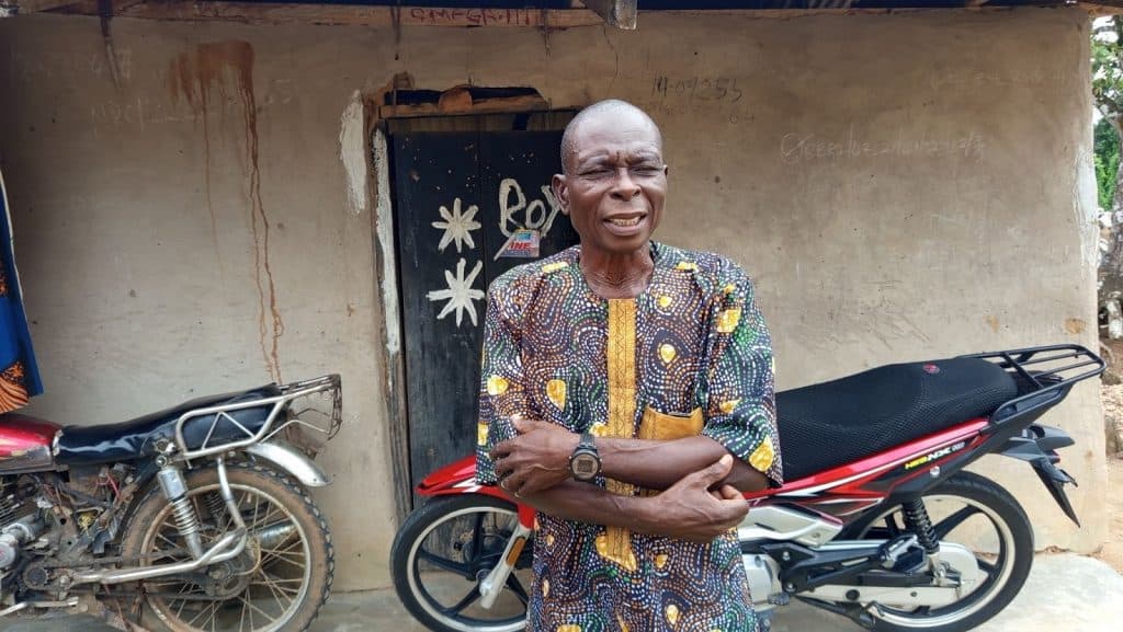 Elderly man in patterned dress stands outside a rustic building beside a motorcycle, with arms crossed and a gentle smile.