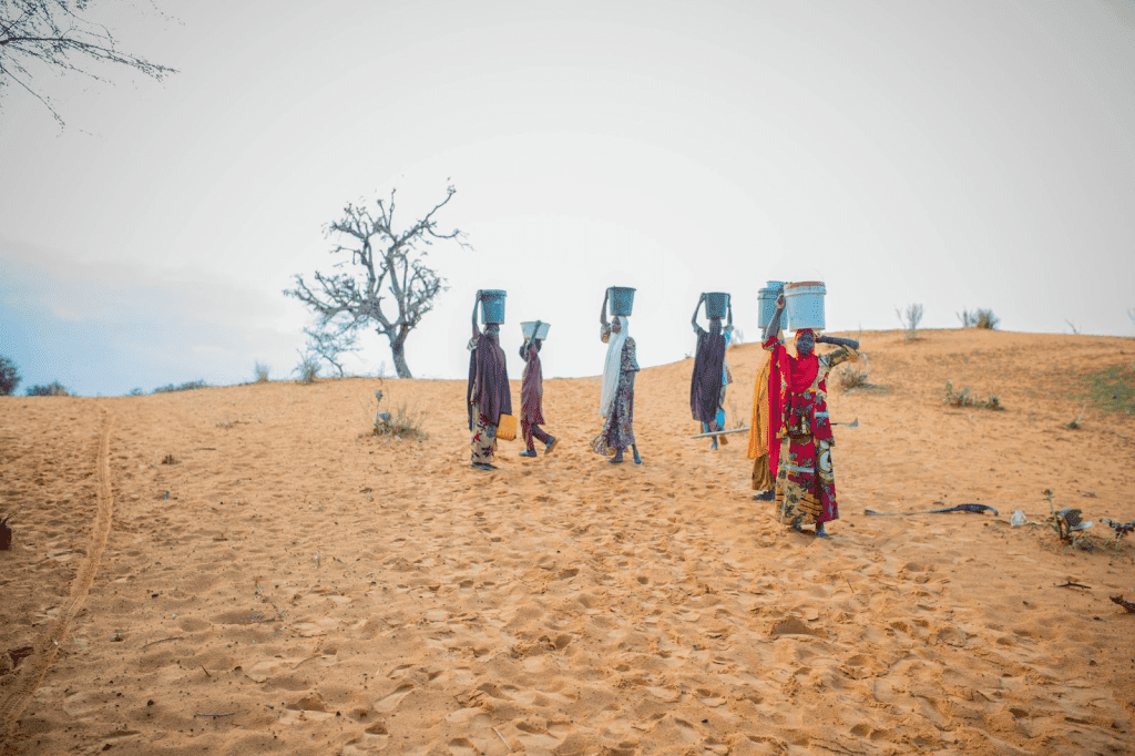 People in colorful clothing walking across sandy terrain, balancing water containers on their heads under a clear sky.