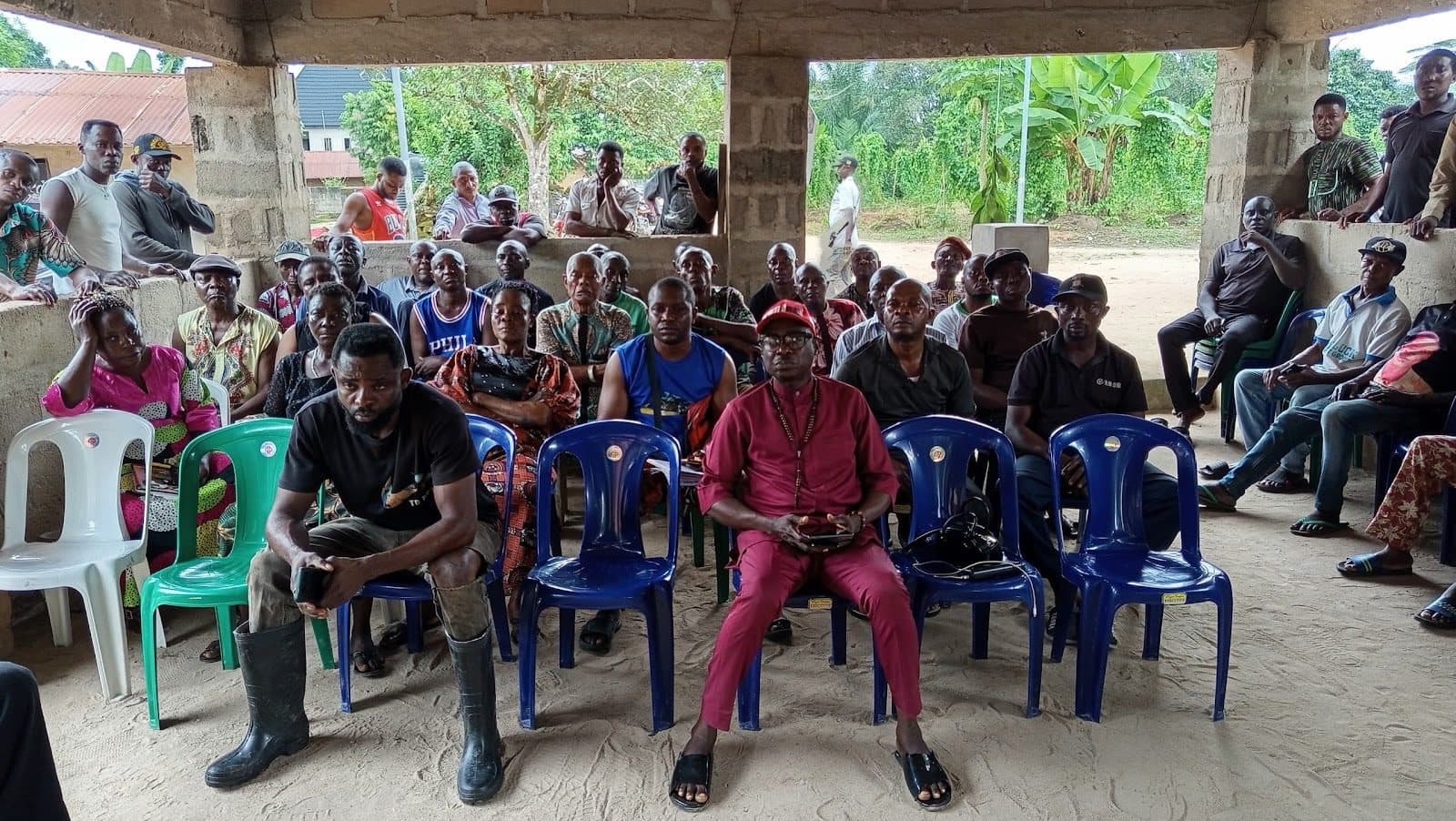 A group of people seated and standing under a shelter, attentively facing forward in an outdoor community gathering.