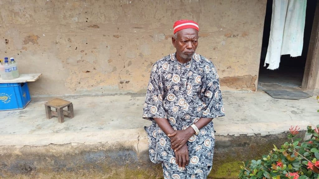 Elderly man in patterned attire and red cap sitting on a low wall outside a rustic building with a small bench and bottles nearby.