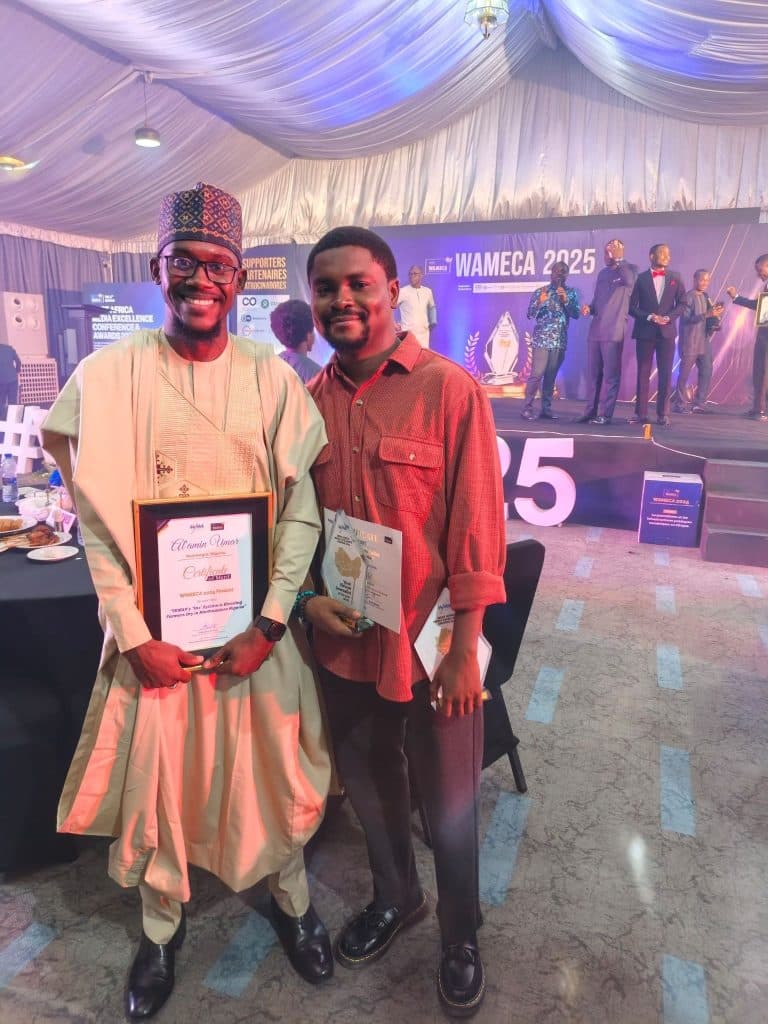 Two men smiling, holding certificates at the WAMECA 2023 event, with a stage and other attendees in the background.