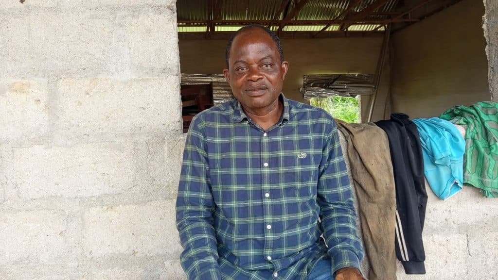 Man in a checkered shirt sits on a concrete wall inside a rustic building, with jackets and shirts draped beside him.