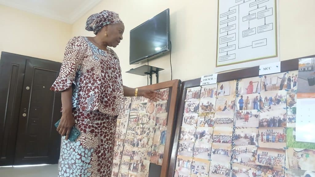 A woman in patterned attire points at a collage of photos on a wall, standing near a TV and a door in an indoor setting.