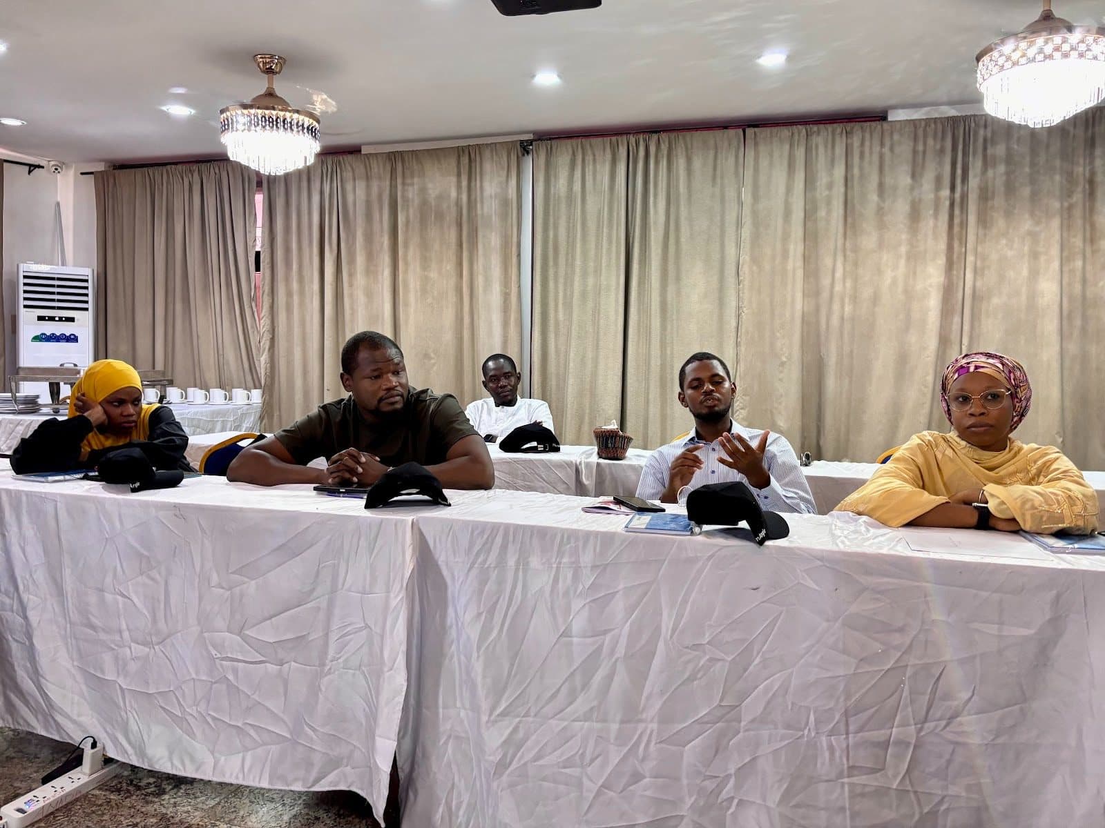 Five people seated at a conference table in a room with chandeliers and curtains.