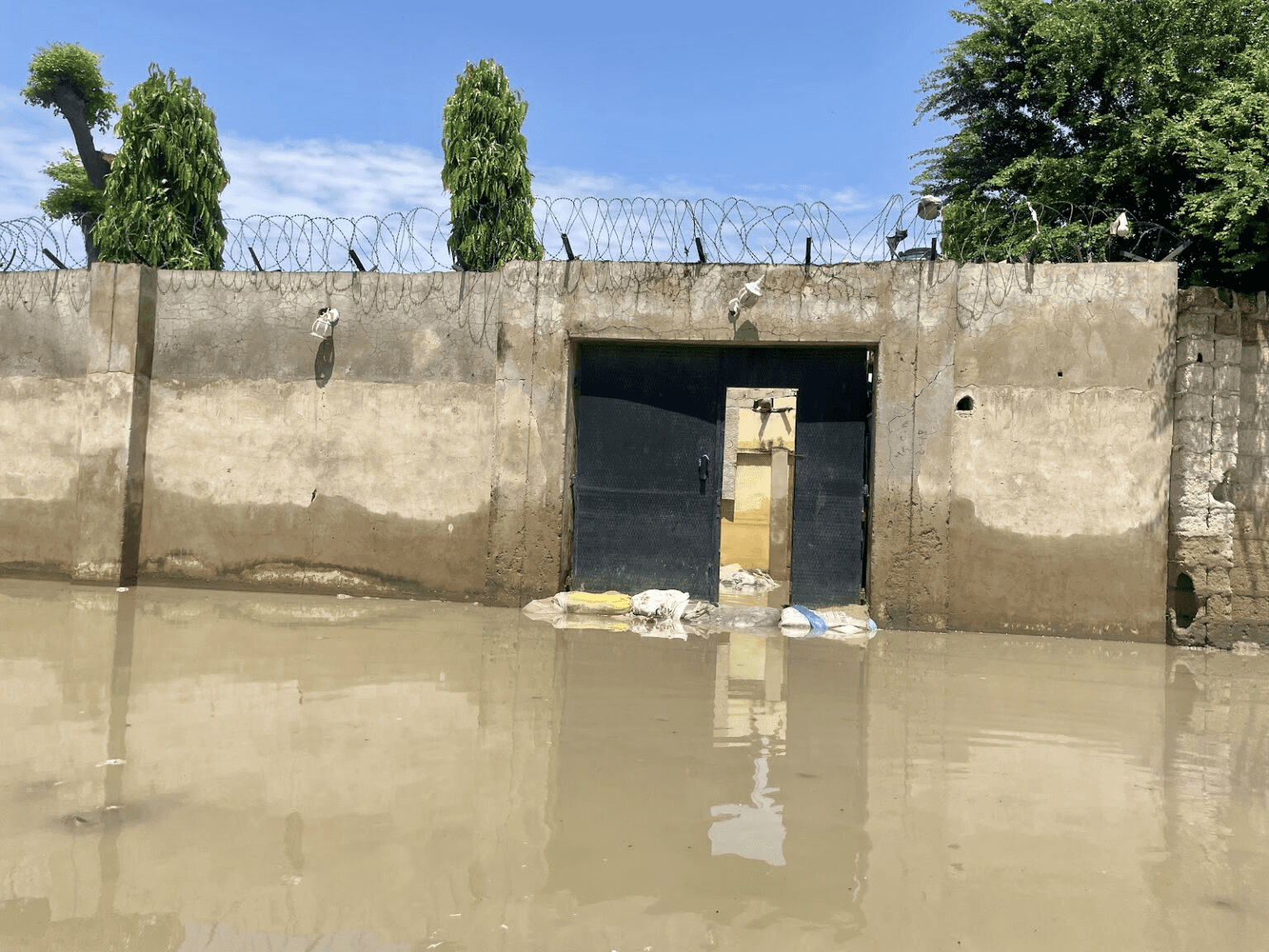 Flooded entrance with sandbags, barbed wire-topped wall, and trees against a clear sky.