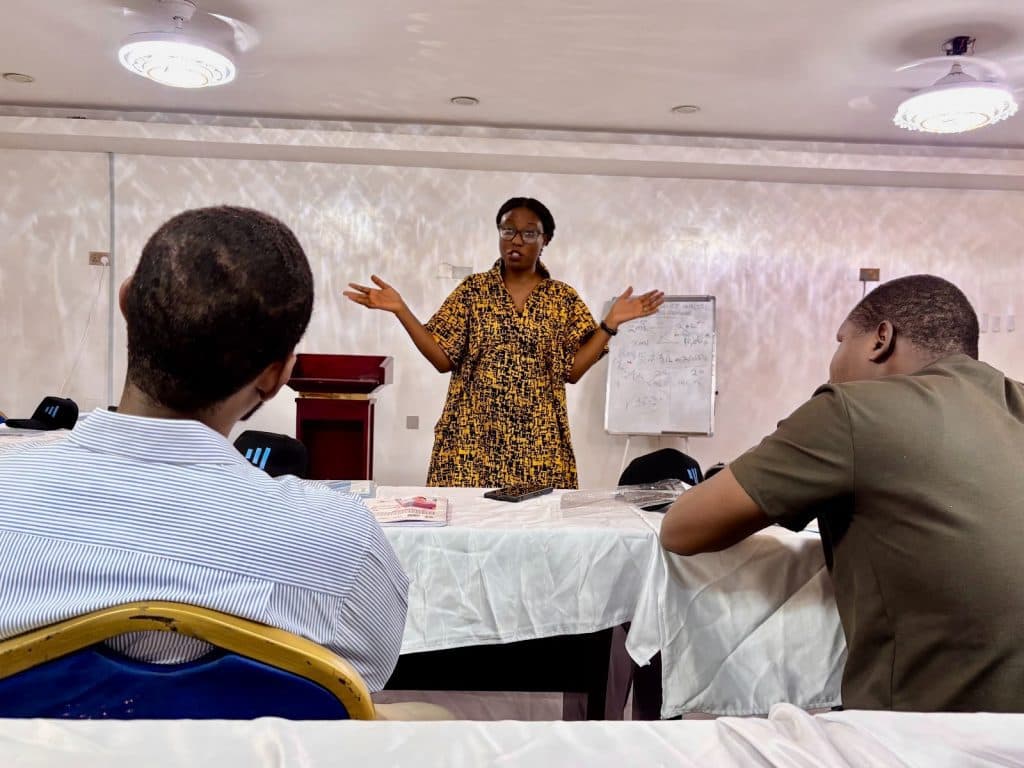 A person stands in front of a group giving a presentation, with a whiteboard and lectern in the background.