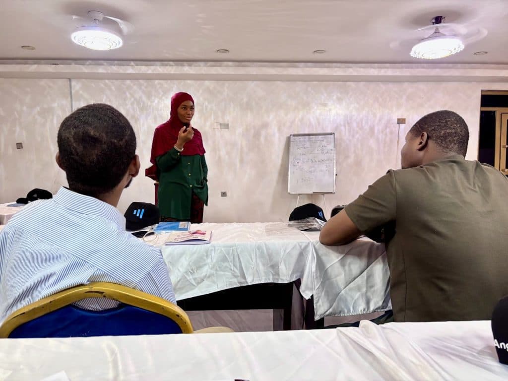 A woman in a red hijab speaks to two seated men in a meeting room, with notes on a flipchart in the background.