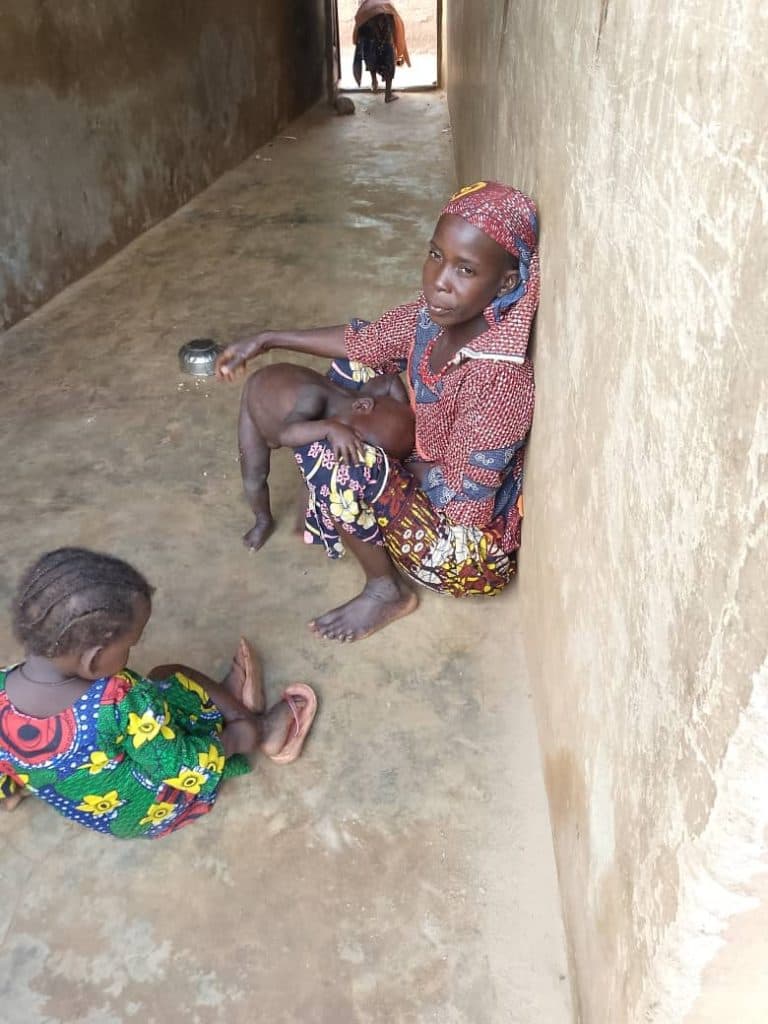 A woman sitting in a corridor holding a baby, with another child nearby. A person is in the background.