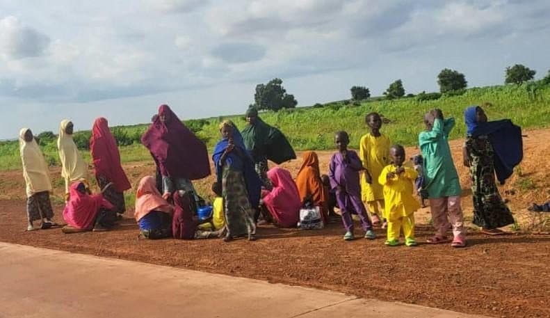 A group of people in colorful clothing standing and sitting on a dirt path with green fields and trees in the background under a cloudy sky.