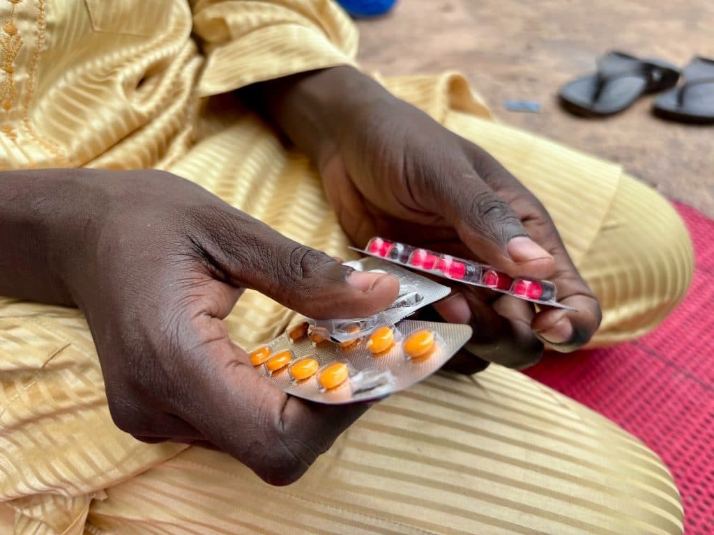 Hands holding blister packs of red and orange pills, with sandals on the ground nearby.