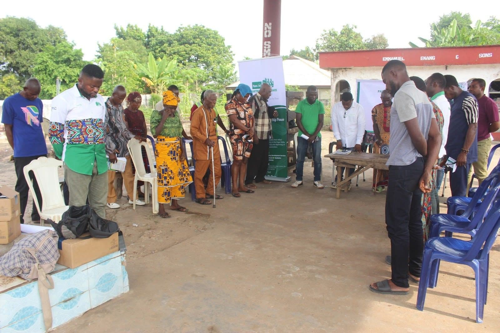 A group of people standing in a semicircle outdoors, some bowing their heads. Boxes and chairs are near them.