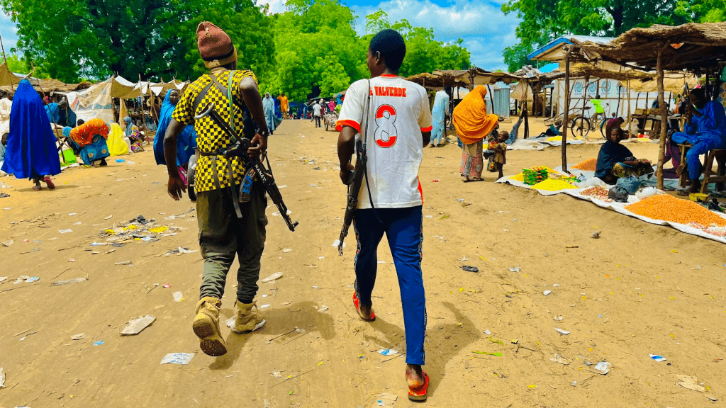 People walk through a vibrant, bustling outdoor market, some carrying rifles; stalls with colorful goods line the street.