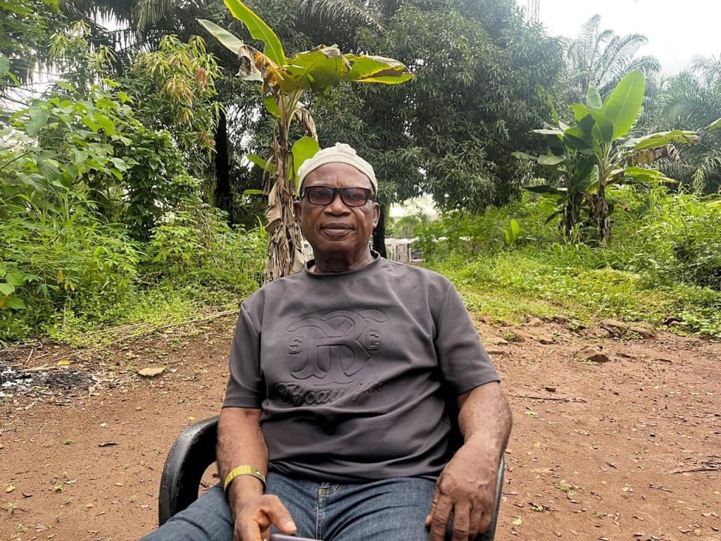 Man sitting outdoors on a chair, wearing a gray shirt and glasses, surrounded by lush greenery and trees.