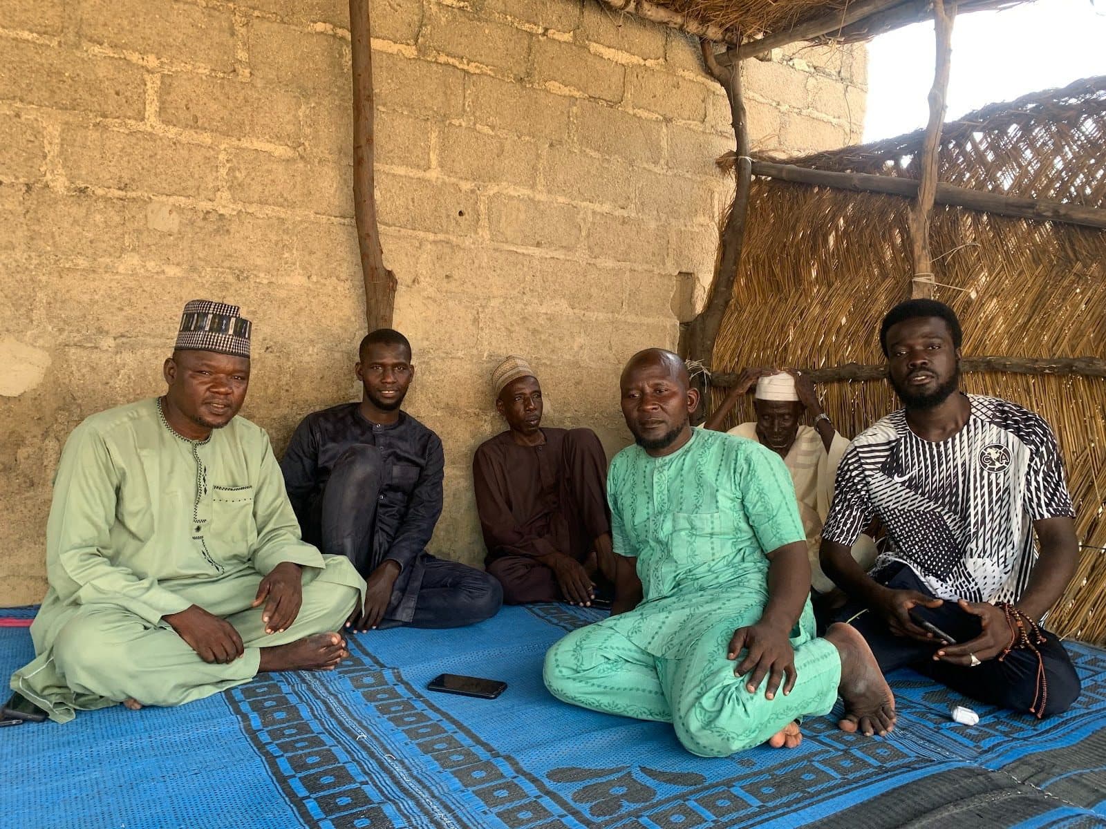 A group of men sitting on a blue mat inside a rustic shelter with straw walls, engaged in conversation.