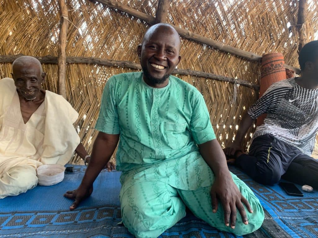 Three men sitting on a blue mat inside a hut with a woven straw wall. They are smiling and wearing traditional clothing.