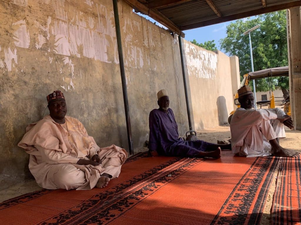 Three men in traditional attire sit on a patterned mat under a shelter, with a wall and trees in the background.