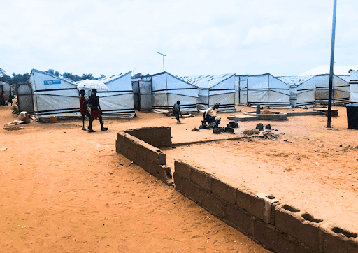 Individuals walk through a refugee camp with white tents on red sandy ground under a cloudy sky.