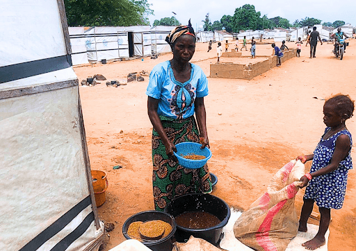 Woman and child sorting grains in a camp setting with temporary shelters in the background.
