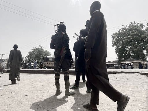 A group of people walking on a sunny day; one person carries a rifle. Trees and buildings are visible in the background.