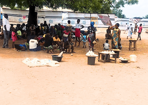 People gather under trees near tents in an outdoor setting, with food items displayed on small tables.