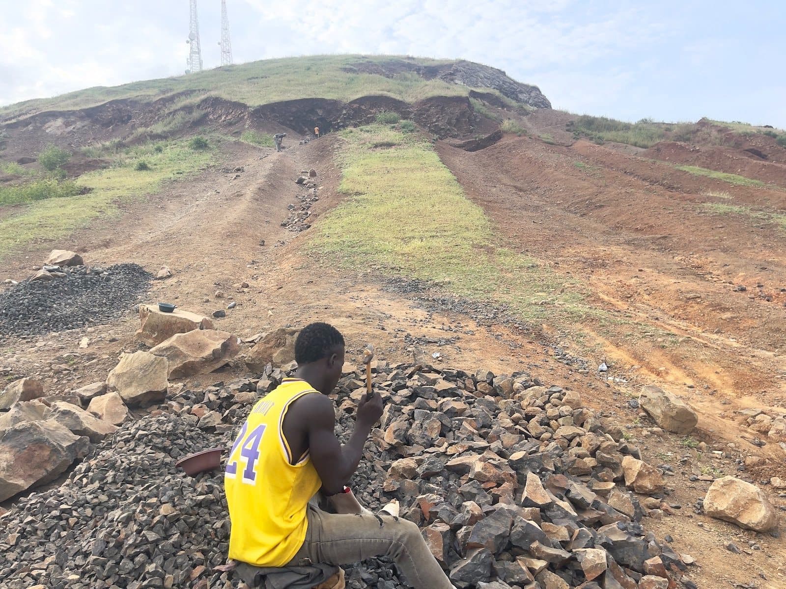 Person in a yellow jersey sitting on rocks with a hill and telecommunications towers in the background under a cloudy sky.