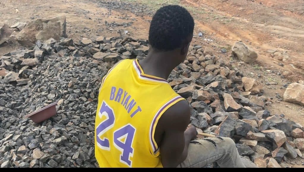 Person wearing a yellow jersey with "Bryant 24" sits on a pile of rocks outdoors.
