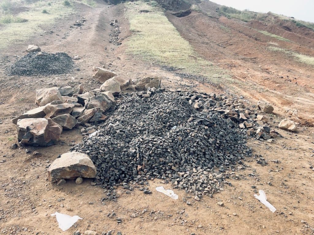 Pile of gravel and rocks on a dirt road with patches of grass in a hilly area.
