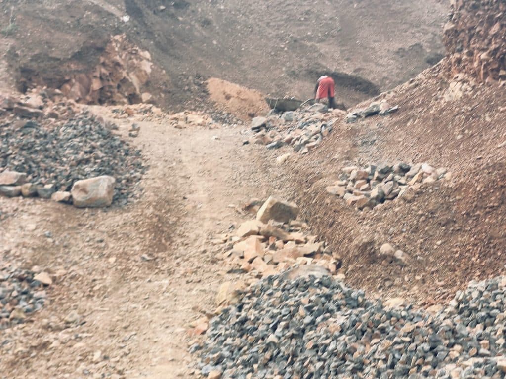 A person in a red shirt stands on a rocky path surrounded by piles of stones in a rugged, uneven terrain.