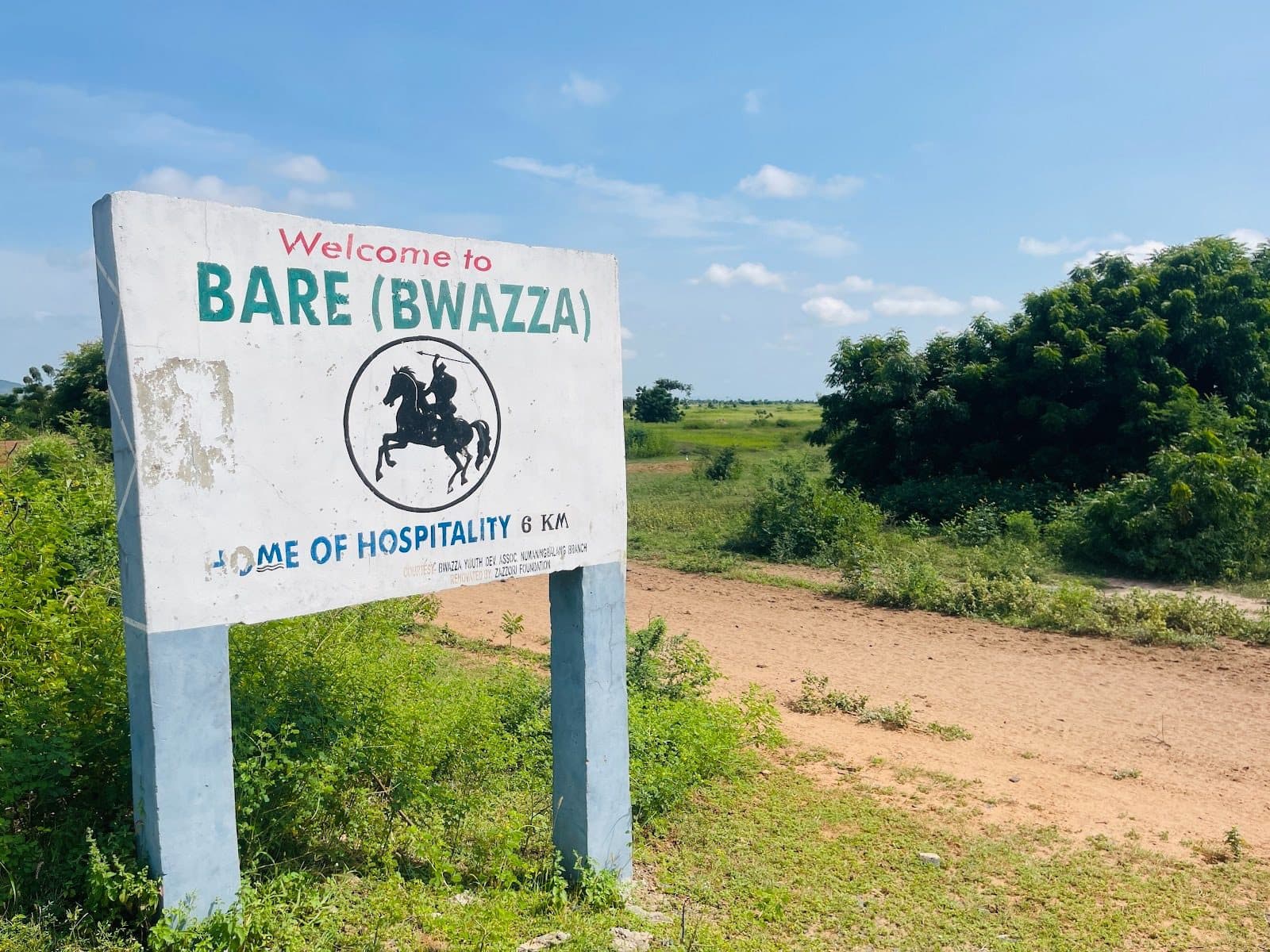 Sign for Bare (Bwazza), "Home of Hospitality," amid greenery under a blue sky. Dirt path leading into the distance.