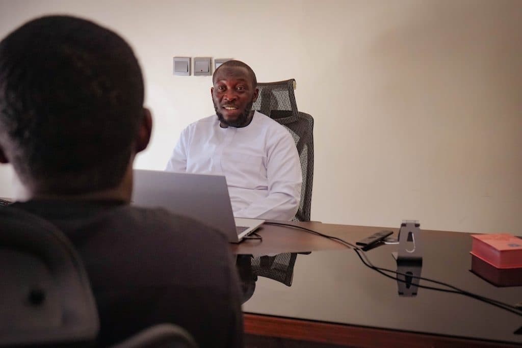 Two people engaged in a conversation in an office setting. One is seated at a desk with a laptop in front.