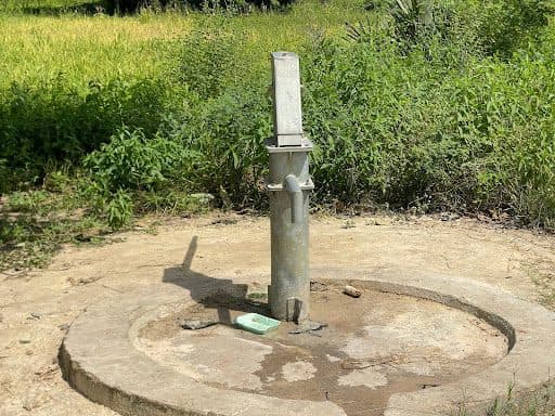 A hand-pump well stands on a concrete base surrounded by green grass and foliage.