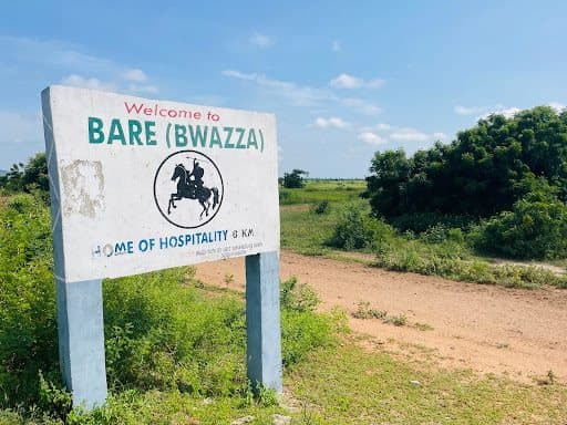 Sign reading "Welcome to Bare (Bwazza), Home of Hospitality," against a backdrop of greenery and blue sky.