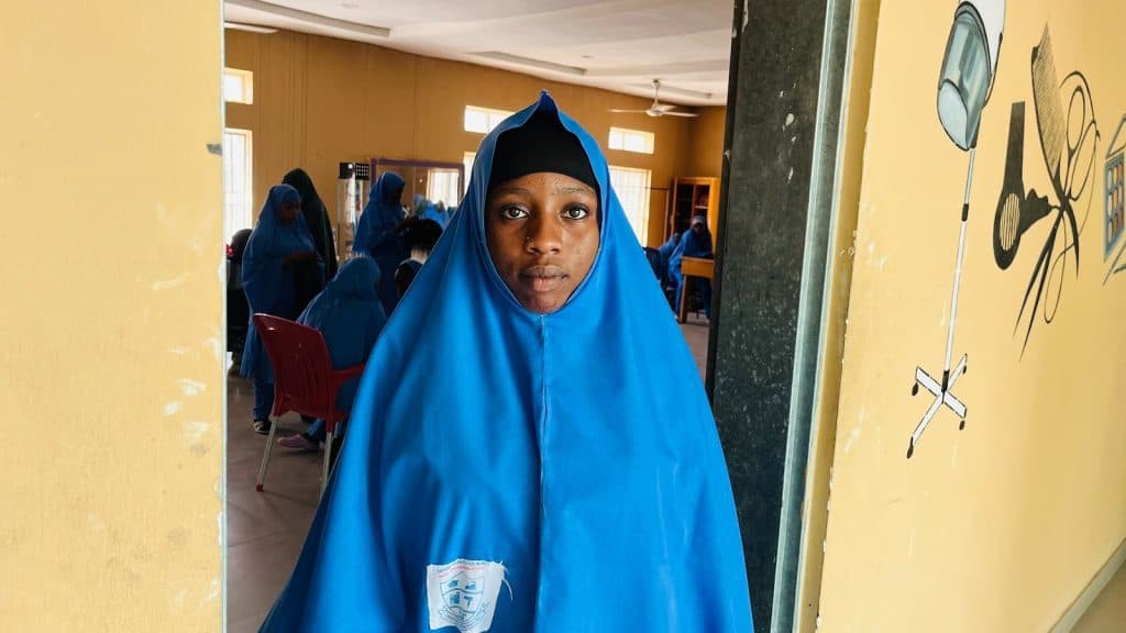 Young person in a blue hijab stands in a room with others in similar attire, with educational wall art visible.