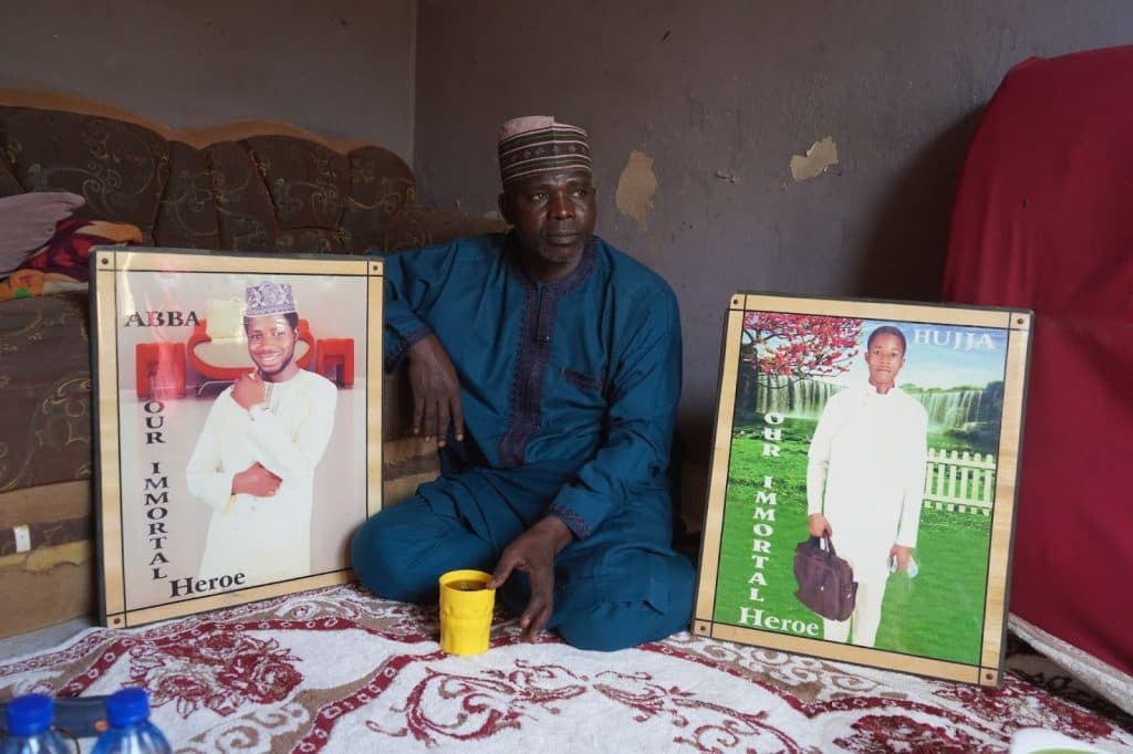 A man in traditional attire sits between two framed portraits labeled "Our Immortal Heroe," holding a yellow cup.