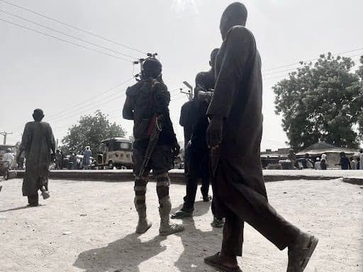 A group of people, including a person equipped with gear, walking in a sunlit area with trees and vehicles in the background.