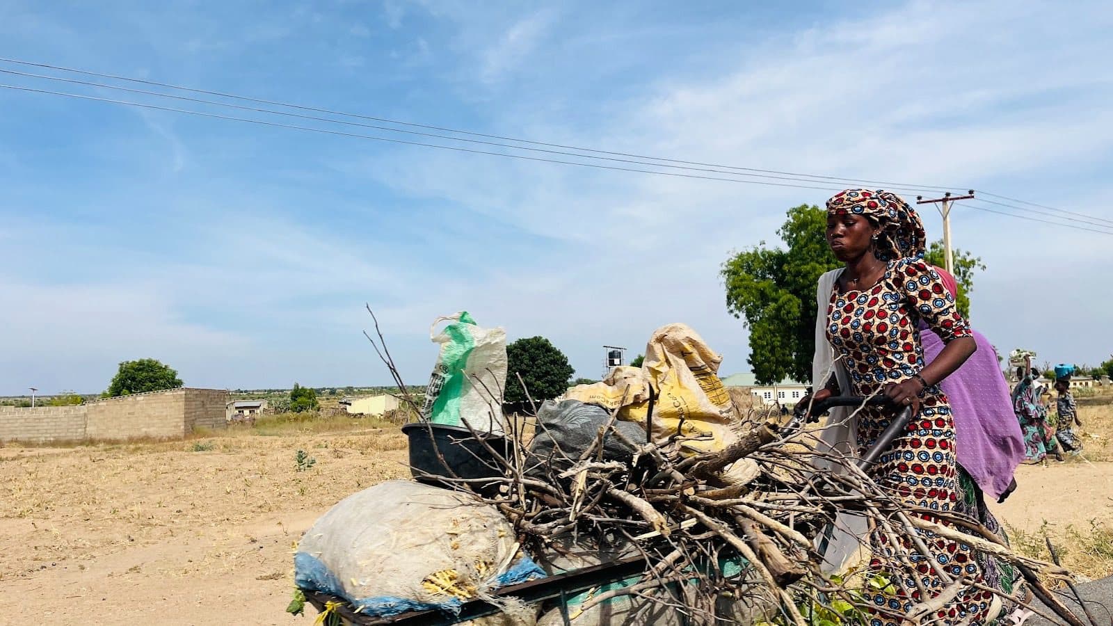 A woman in patterned attire pushes a cart filled with branches and bags along a dirt road, under a clear blue sky.
