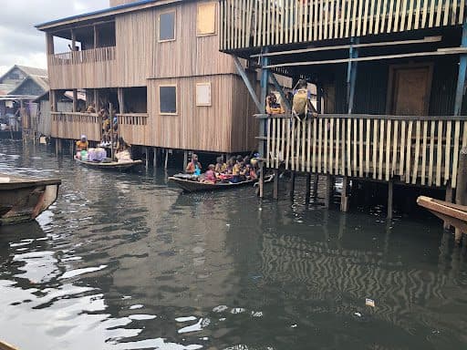 Wooden stilt houses above water with people in boats nearby, navigating a canal-like setting.