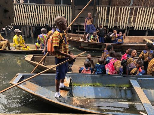 Children in colorful uniforms travel on boats through a canal, surrounded by wooden buildings and other passengers.
