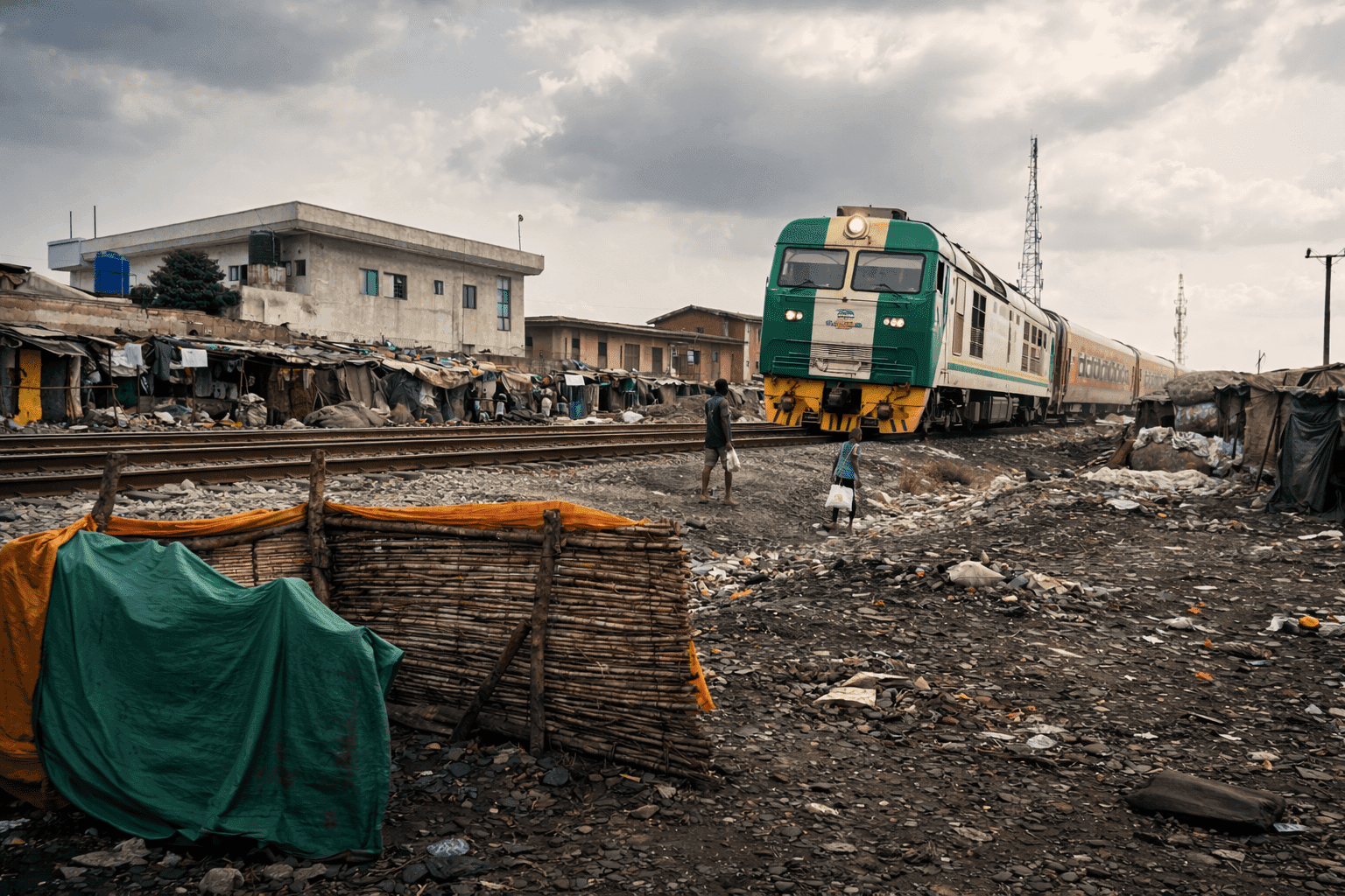 Green train passing through a rundown area with makeshift houses, as two people walk beside the tracks under a cloudy sky.