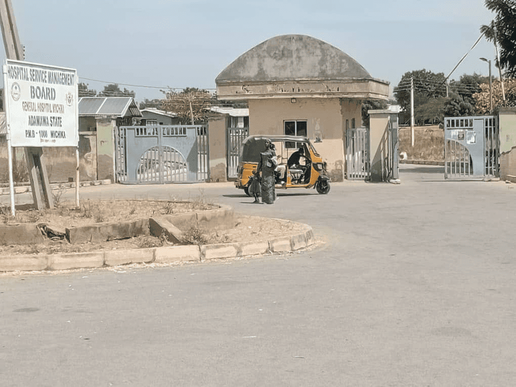 A person in a yellow auto rickshaw outside a hospital gate in Adamawa State, Nigeria, next to a sign for the General Hospital Michika.