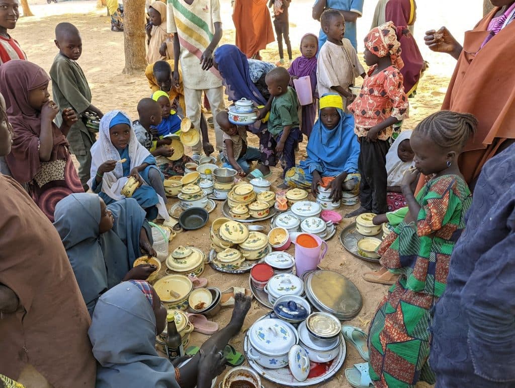 Children gather around a collection of colorful pots and plates, sharing food outdoors.