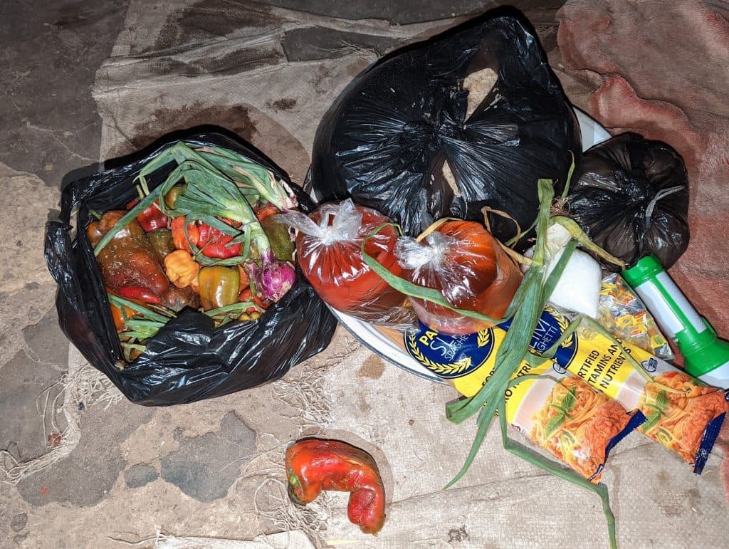 Assorted vegetables, spices, and packaged food items in black bags on a straw mat; includes peppers, spring onions, pasta, and seasoning cubes.