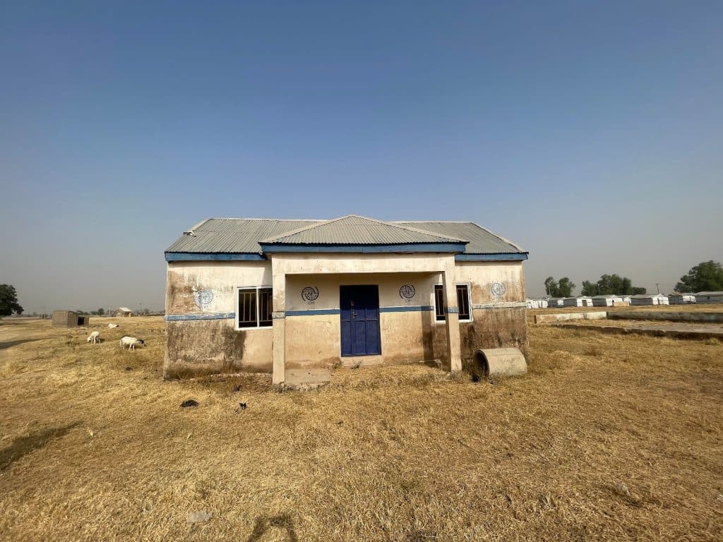 Small, weathered building with a blue door in a dry, grassy field under a clear blue sky. Sheep graze nearby.
